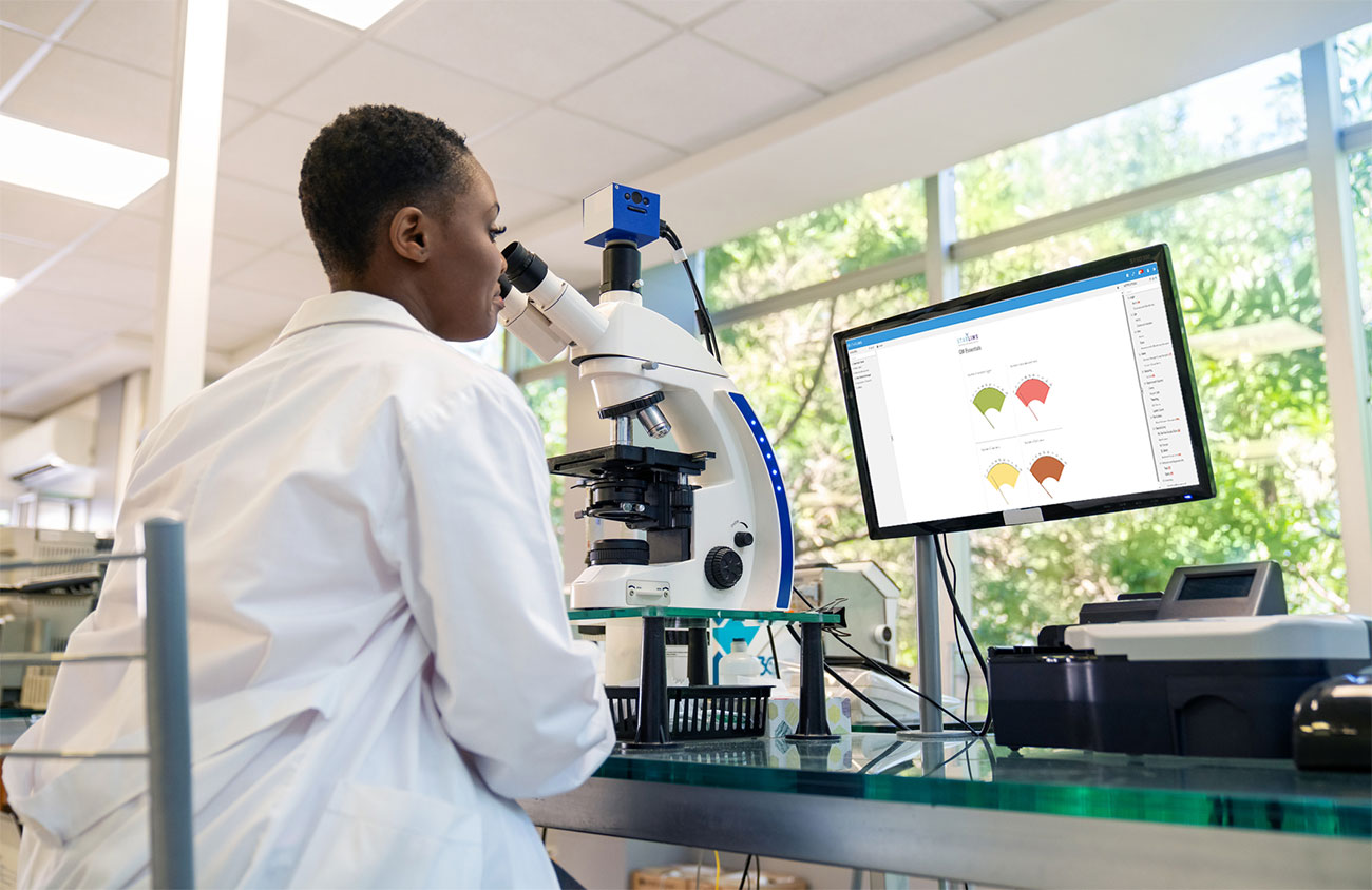 Scientist using a microscope with data displayed on a nearby computer screen