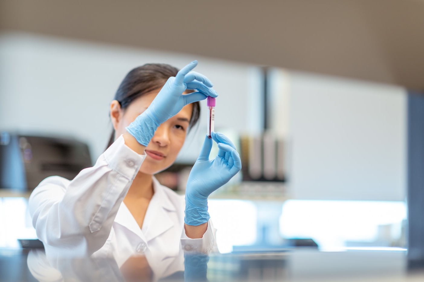A female chemist is working solo in lab. The woman is holding up a small vial of blood and is analyzing the medical sample. She is wearing a lab coat and protective gloves.