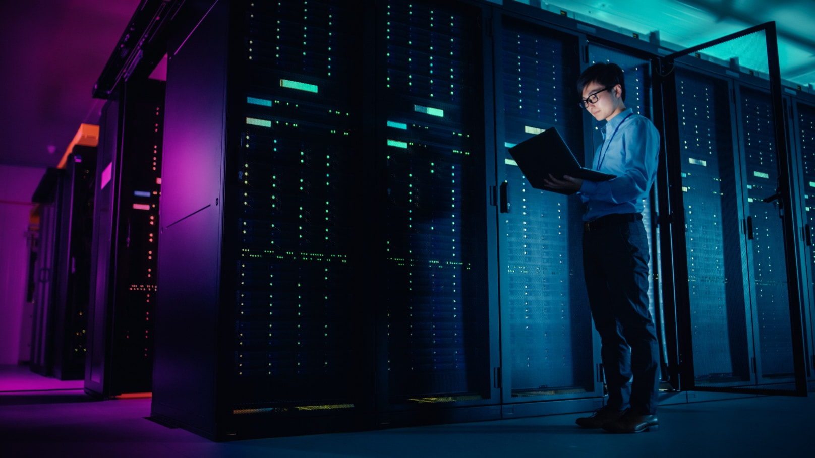 IT professional stands in front of servers with a laptop.