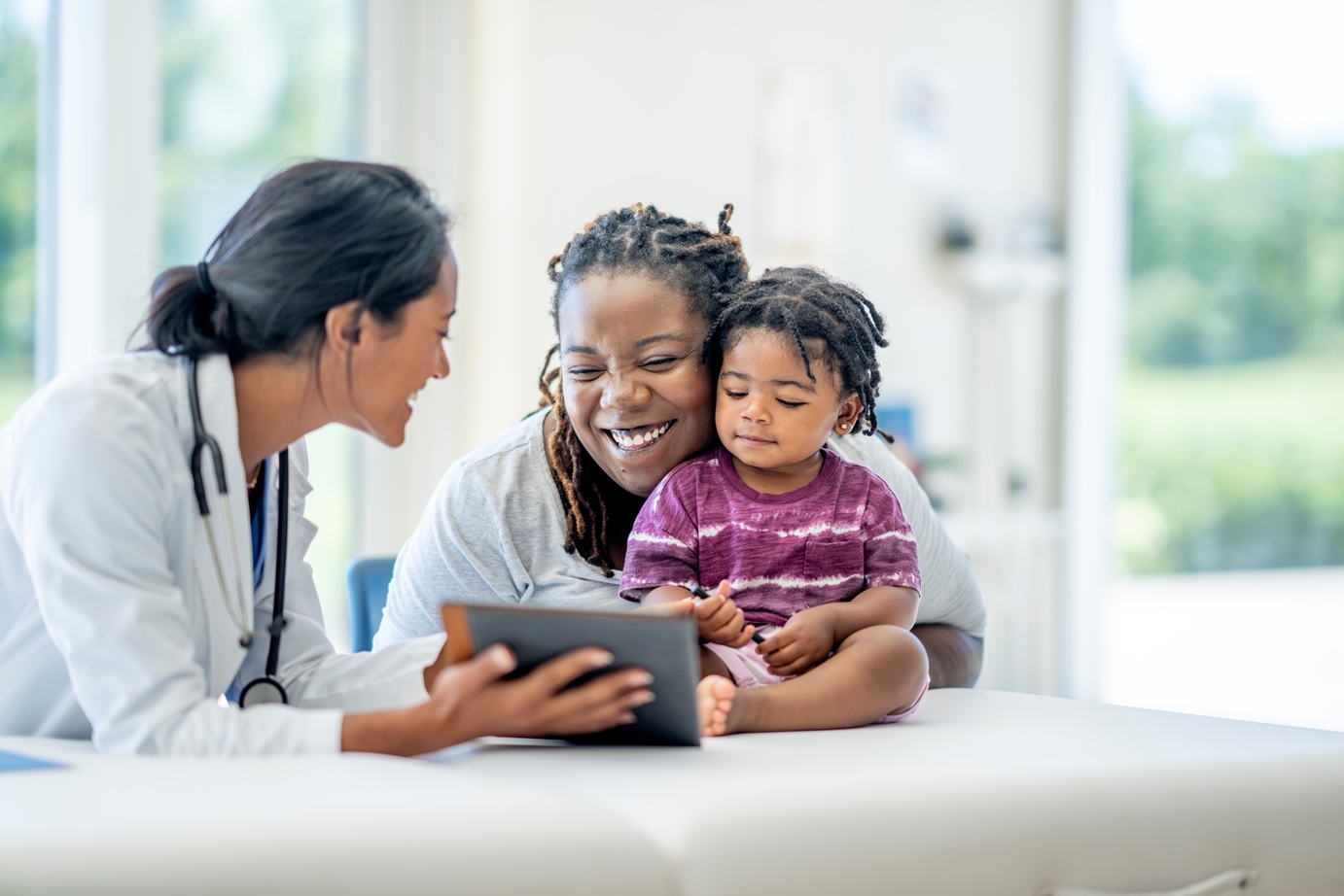 A young Mother and her child visit the doctor together. The baby is seated on the exam table with his mother seated in front of him and making sure he does not fall. Their female doctor is seated in front of them, showing them her clinical lab informatics software on a tablet to them and smiling as they talk.